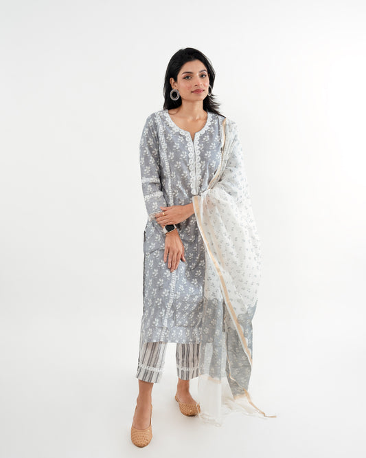 Front View: A model showcasing the Floral Grey with Lace Work Striped Pant Cotton Suit from Cottons Daily. The grey floral-printed kurta features a round V-neck highlighted with a delicate lace border and 3/4th sleeves. Paired with white printed straight-fit pants and a Chanderi white floral print dupatta with a golden border, this ensemble exemplifies elegance and comfort.