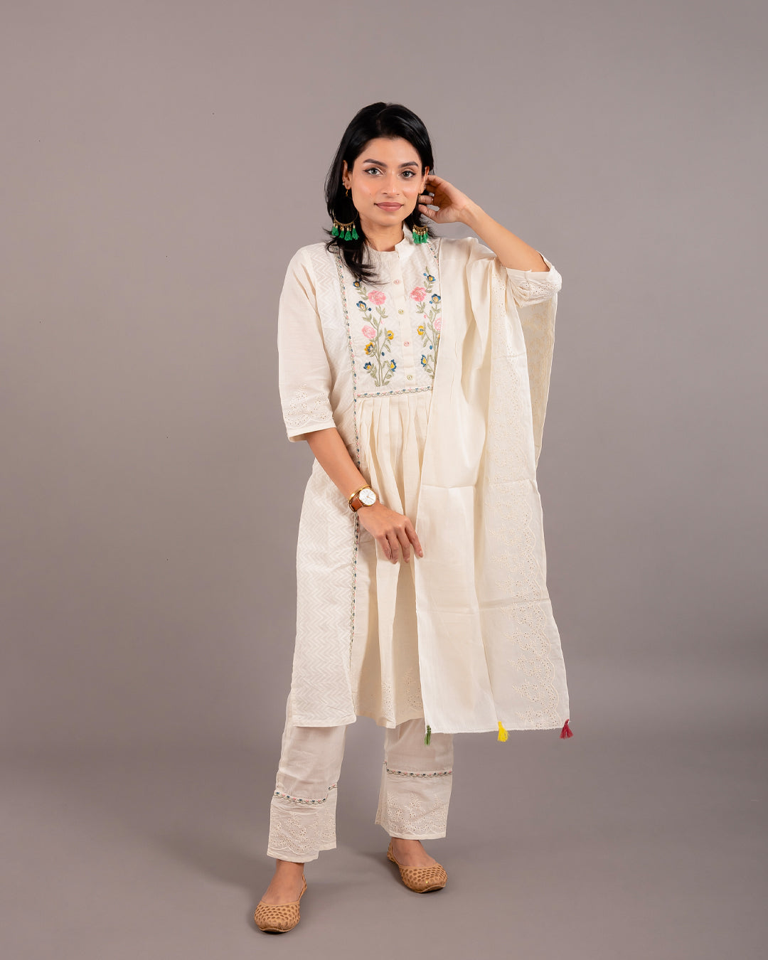 A model wearing an Off White Floral Cotton Suit from Cottons Daily, featuring a French collar kurta with floral embroidered front design and 3/4th sleeves, paired with straight-fit pants highlighted with embroidery work, and accessorized with a cotton stole with tassels.