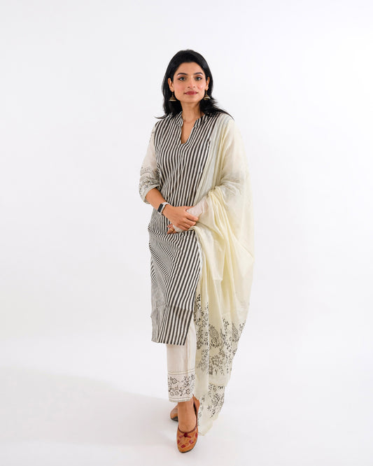 A woman elegantly dressed in a stripe printed black and white cotton suit by Cottonsdaily, featuring vertical stripes on the kurta, paired with a delicate white dupatta adorned with floral embroidery, and complemented by simple white embroidered pants and traditional juttis.